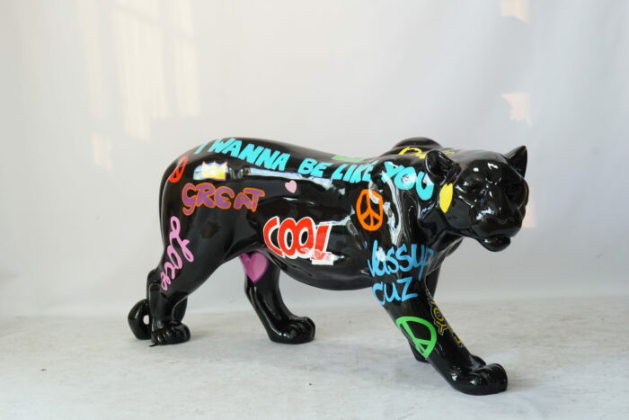 A black panther sculpture with colorful graffiti-style messages and symbols, including phrases like "I WANNA BE LIKE YOU," "COOL," "GREAT," and "wassup cuz," as well as peace signs and hearts.