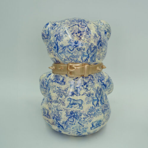 I don't recognize any text in Polish in the provided image. The image shows the back of a ceramic bear figurine with a blue pastoral print pattern and a gold-colored collar.