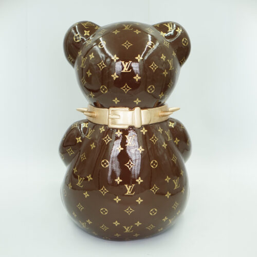 The image shows a brown teddy bear figurine adorned with a gold Louis Vuitton monogram pattern, dressed in a spiked belt.