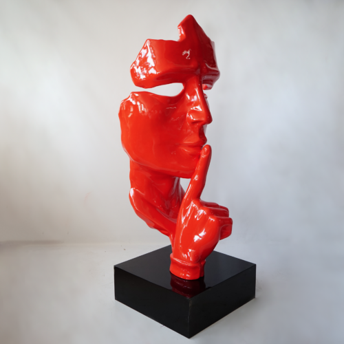 Red abstract sculpture of a face with a finger raised to the lips, suggesting silence, mounted on a black base.