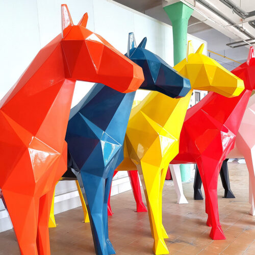 Geometric, large, colorful horse sculptures in a row, each in a different vibrant color including orange, blue, yellow, red, and white, displayed indoors.