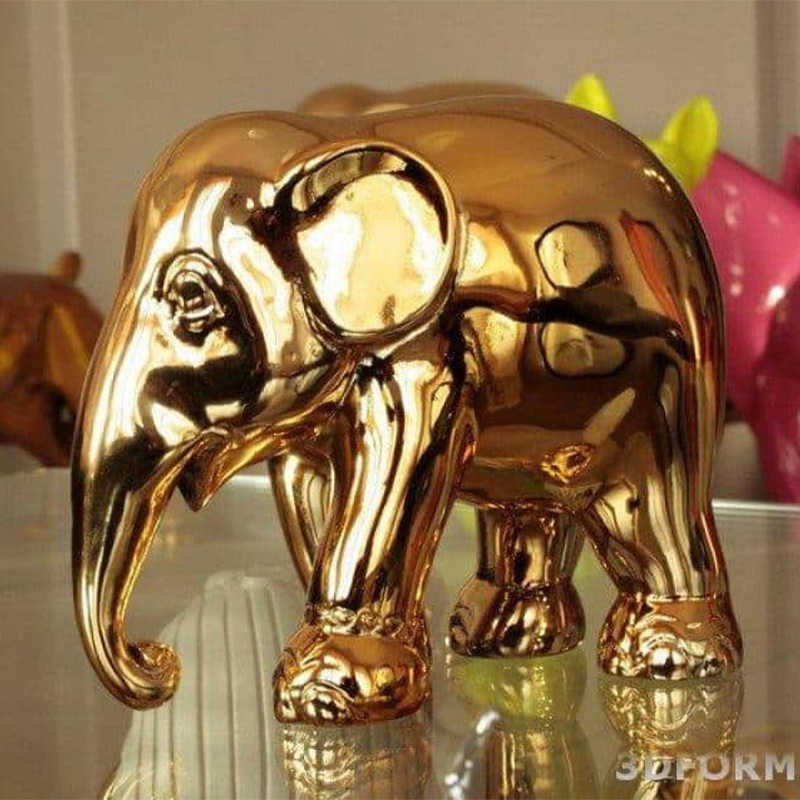 A shiny golden elephant figurine displayed on a reflective surface with colorful objects in the background.