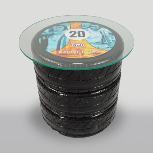 A table made from four stacked racing tires with a glass top featuring a design that includes the Gulf logo, the number 20, and images of two race car drivers.
