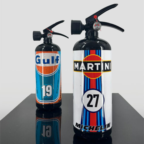 Two fire extinguishers with custom racing liveries, one with Gulf branding and the number 19, and the other with Martini and Michelin branding and the number 27, placed on a reflective surface.