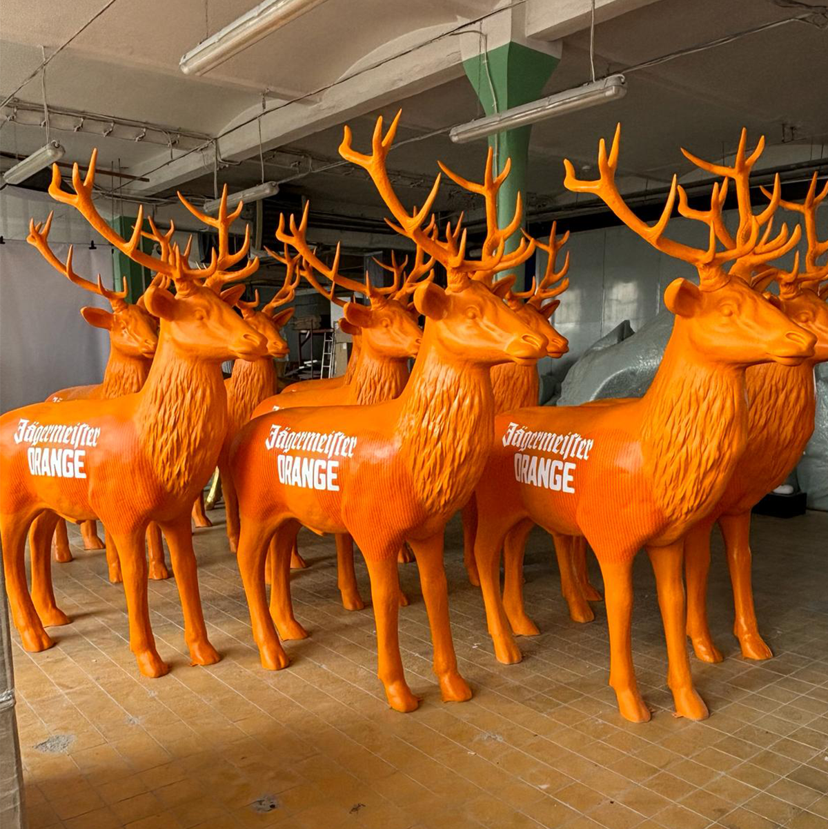 Several orange statues of deer with large antlers, displaying the text "Jägermeister ORANGE" on their sides, are standing in a warehouse.