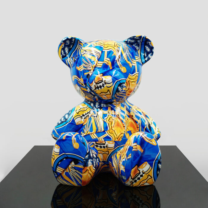 Sculpture of a bear decorated with a colorful pattern, primarily blue with yellow and orange accents, seated on a black reflective surface.