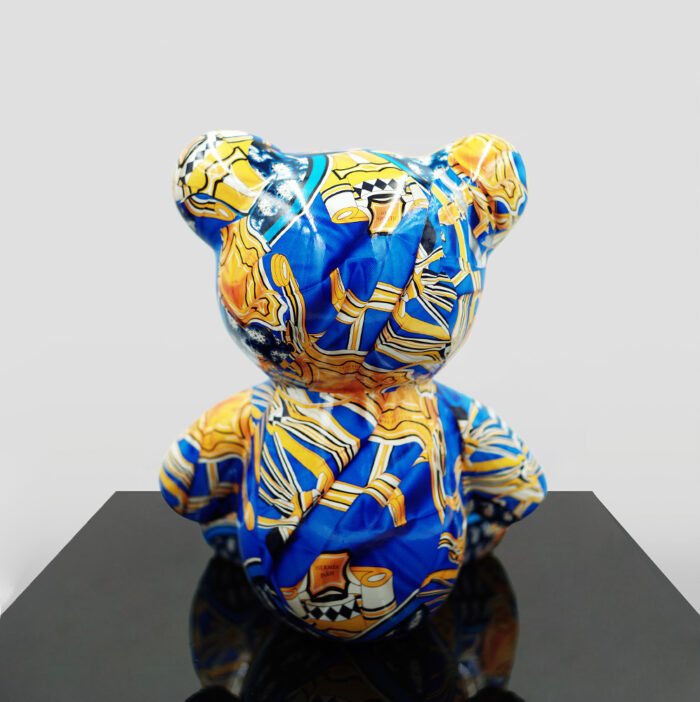 A teddy bear figure wrapped in a colorful printed fabric with blue, yellow, and white patterns, displayed on a dark reflective surface.