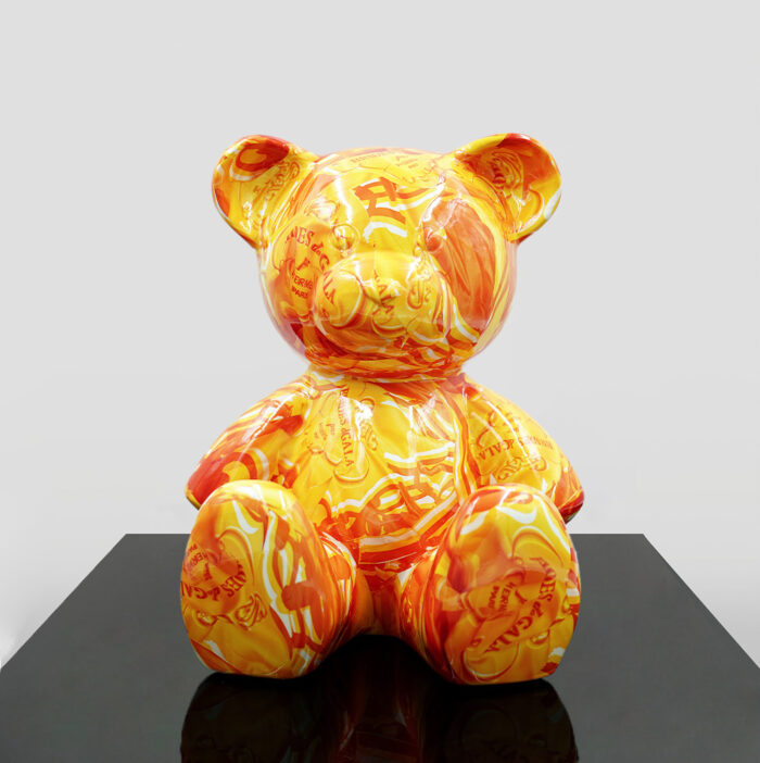 A large, stylized teddy bear sculpture with a vibrant orange and yellow pattern featuring various text and images. The sculpture sits on a glossy black surface against a plain white background.