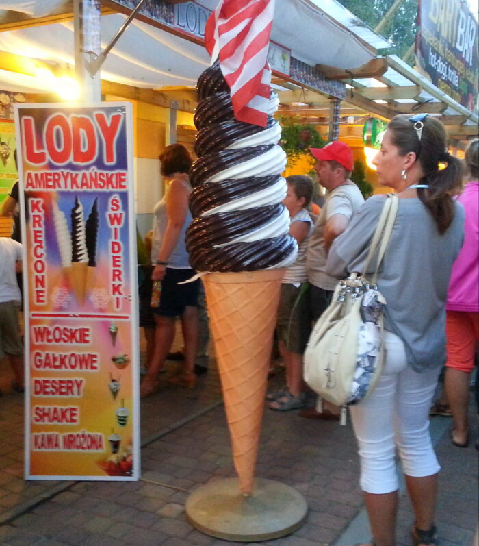 Large cone-shaped ice cream statue with chocolate and vanilla swirls, near a stand selling various ice cream products, with people queuing. Sign features "Lody Amerykańskie" and other Polish text.