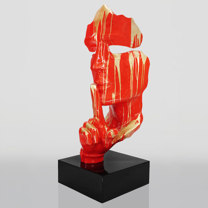 Abstract sculpture of a red and gold face with an elongated finger raised to closed lips, mounted on a black square base.