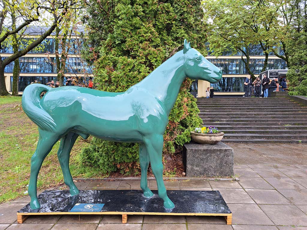 oryg_064490685_1129949550544944_1465385417682452480_o Green horse statue on a wooden platform, placed on paved ground, surrounded by trees and a building with steps in the background.