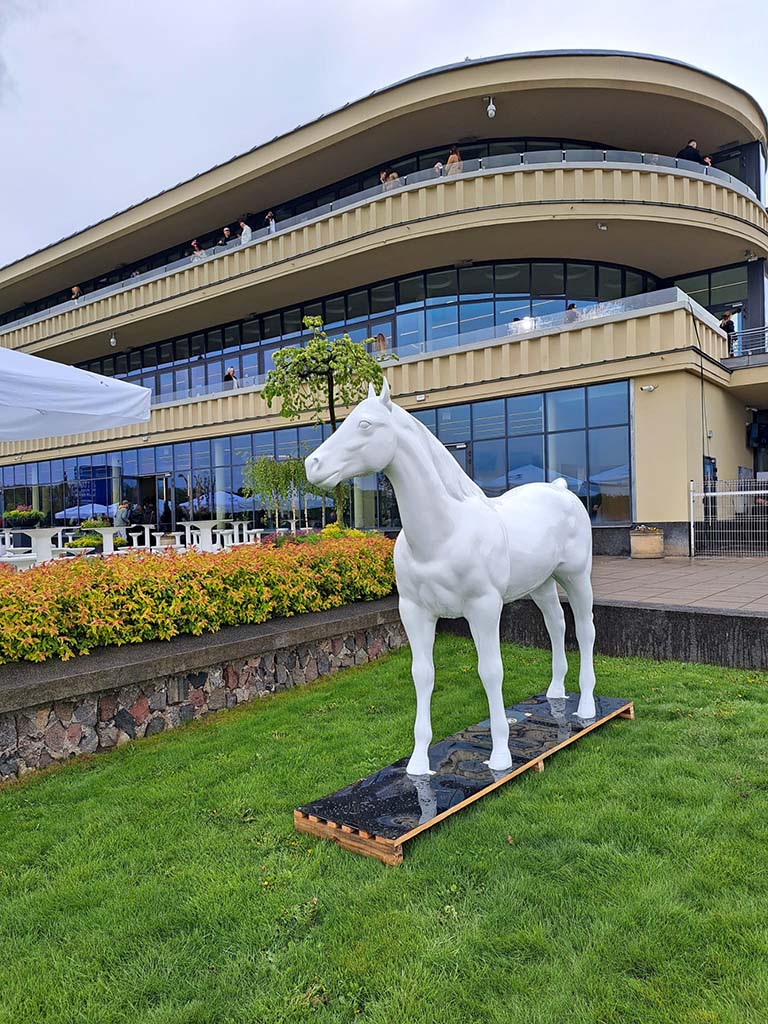 Colorful Horse Sculptures at the Partynice Racecourse in Warsaw 1 Biały pomnik konia na trawniku przed nowoczesnym budynkiem z dużymi oknami.