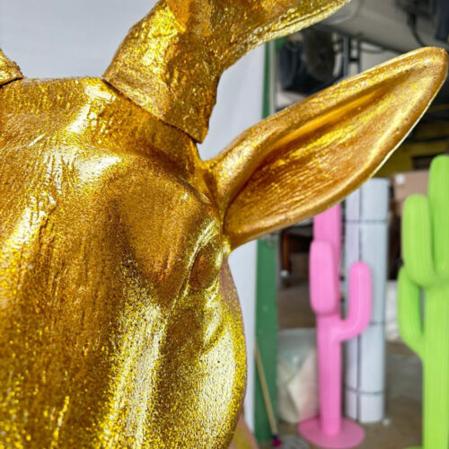 Close-up of a golden animal sculpture with visible ears, and out-of-focus colorful cactus-shaped objects in the background.