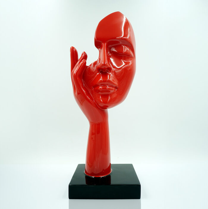 Red abstract face sculpture resting on a black base with a hand mimicking a contemplative pose.