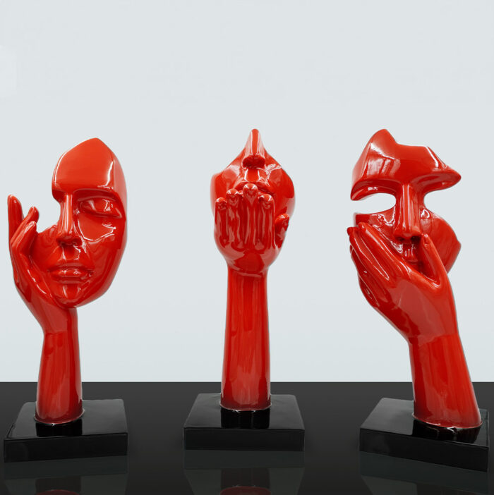 Three red abstract sculptures of human faces, each supported by a single hand, against a plain background.