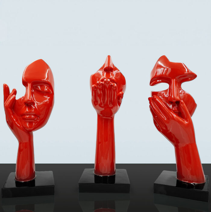 Three red abstract sculptures of human faces supported by hands on black bases.