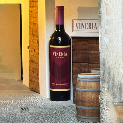 Large decorative wine bottle with a label reading 'Vineria Carin Wine & Restaurant' standing outside a building next to a wooden door and two wooden wine barrels on a cobblestone street.