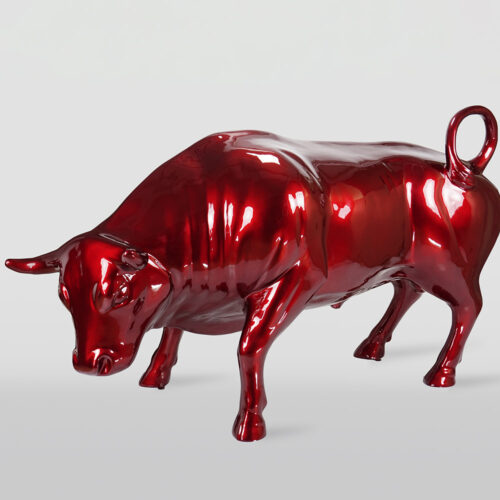 Glossy red sculpture of a bull with its head lowered and tail curled upwards on a light gray background.