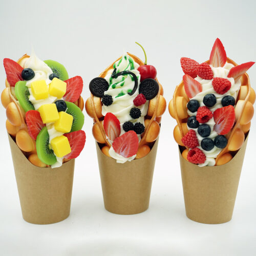 Three bubble waffles in brown paper cups topped with whipped cream and various fruits including strawberries, blueberries, kiwis, mango cubes, raspberries, blackberries, a cherry, and mini chocolate cookies, arranged in a row on a white background.