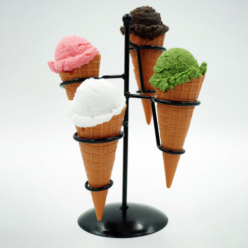 Four ice cream cones with scoops in pink, white, dark brown, and green colors placed in a black metal ice cream cone holder against a white background.