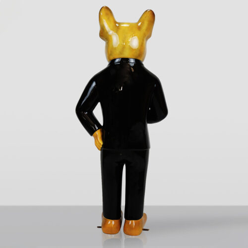 Rear view of a glossy ceramic figurine with a yellow animal head wearing a black suit, standing on a reflective surface against a plain gray background.