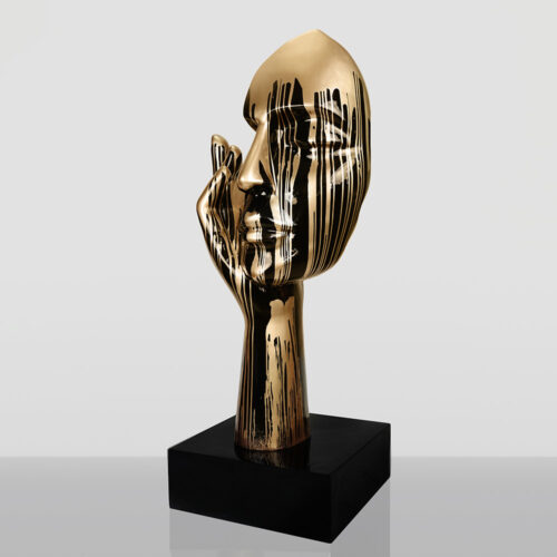 Abstract golden sculpture of a human face resting on a hand with vertical black paint drips, mounted on a black square base.