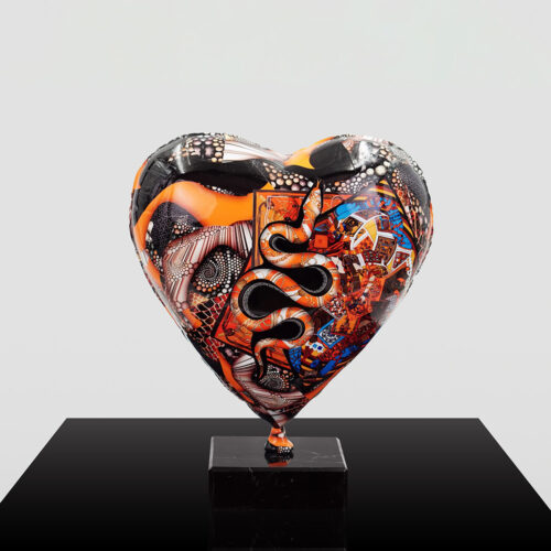 Heart-shaped sculpture with intricate patterns in black, orange, white, and blue, featuring snake and abstract designs, mounted on a black square base against a plain gray background.
