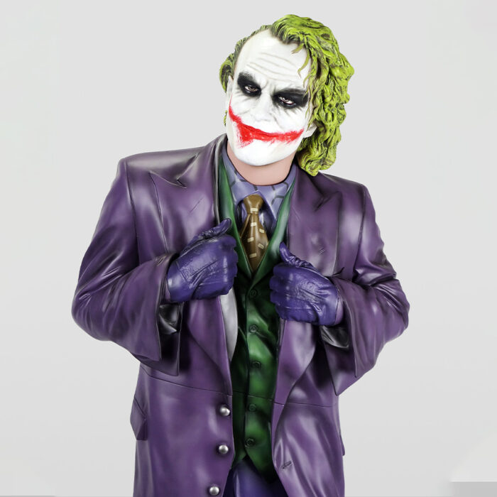 Person dressed as the Joker character, wearing a purple suit jacket, green vest, brown tie, purple gloves, with white face paint, black eye makeup, red painted smile, and green hair, posing against a plain light background.