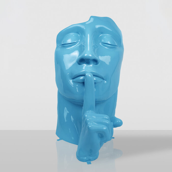Glossy blue sculpture of a face with eyes closed and a finger pressed to the lips in a shushing gesture, placed on a reflective surface with a plain light gray background