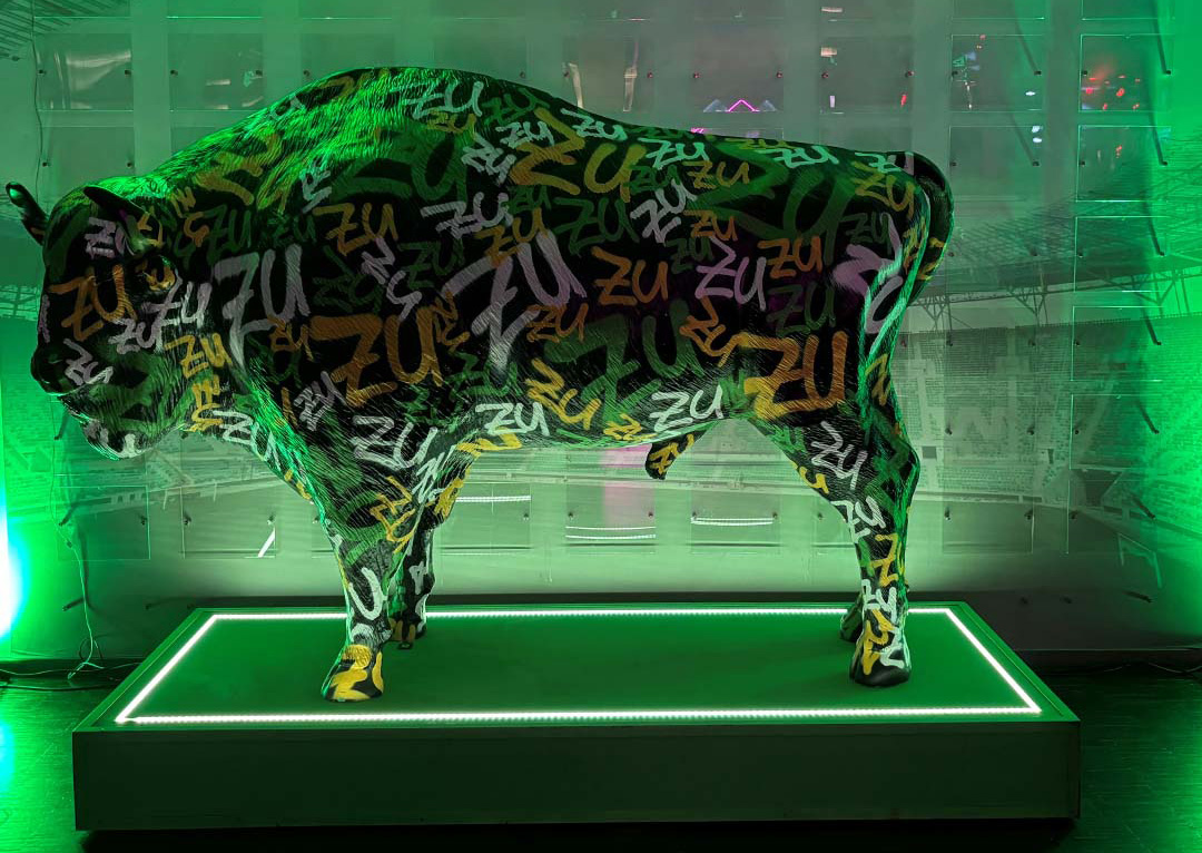Sculpture of a cow covered in graffiti-style text "ZU" in various colors, displayed on a lit platform with green lighting in the background.