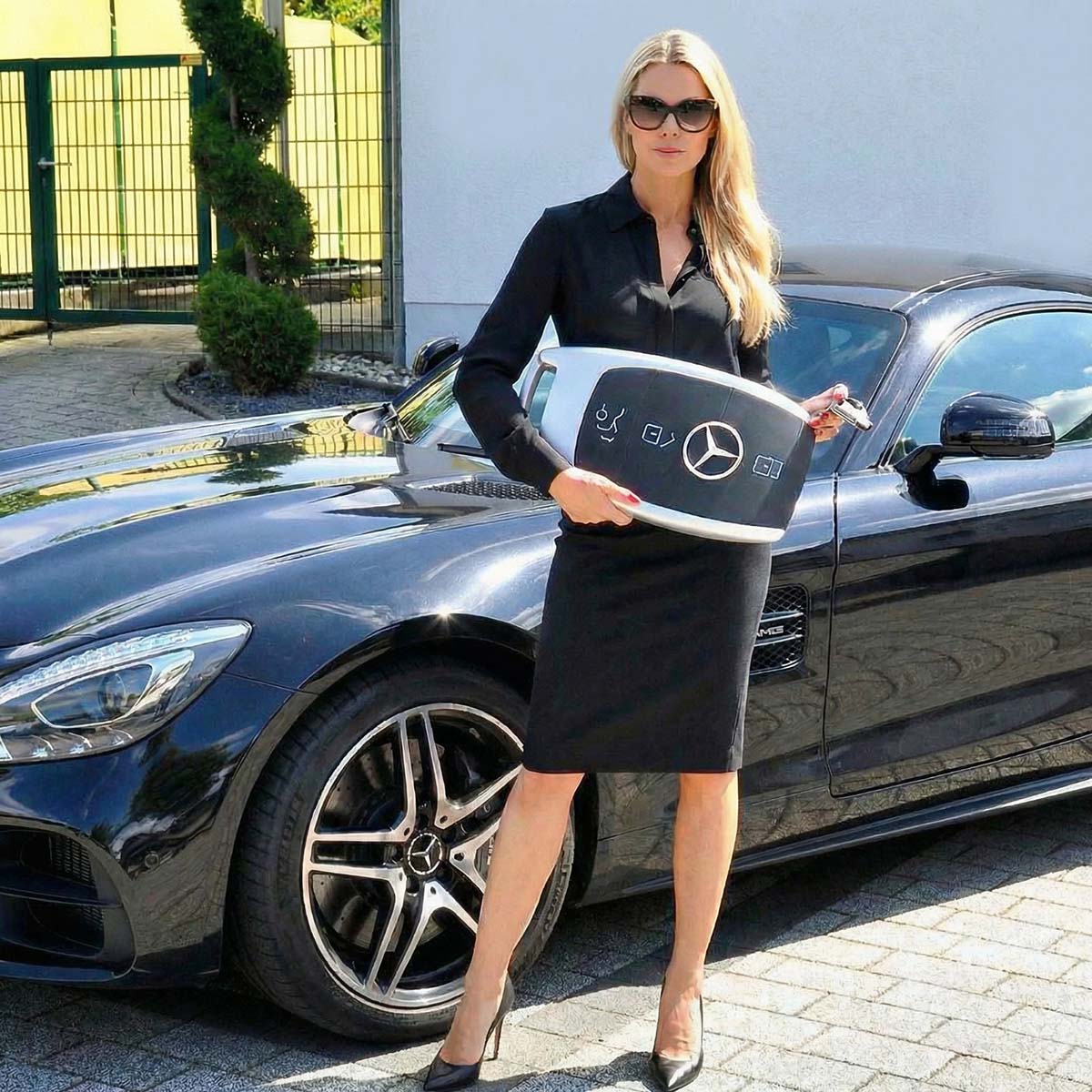 Kobieta w eleganckiej czarnej sukience trzyma dużą figurę reklamową w kształcie kluczyka Mercedes, stojąc obok auta.