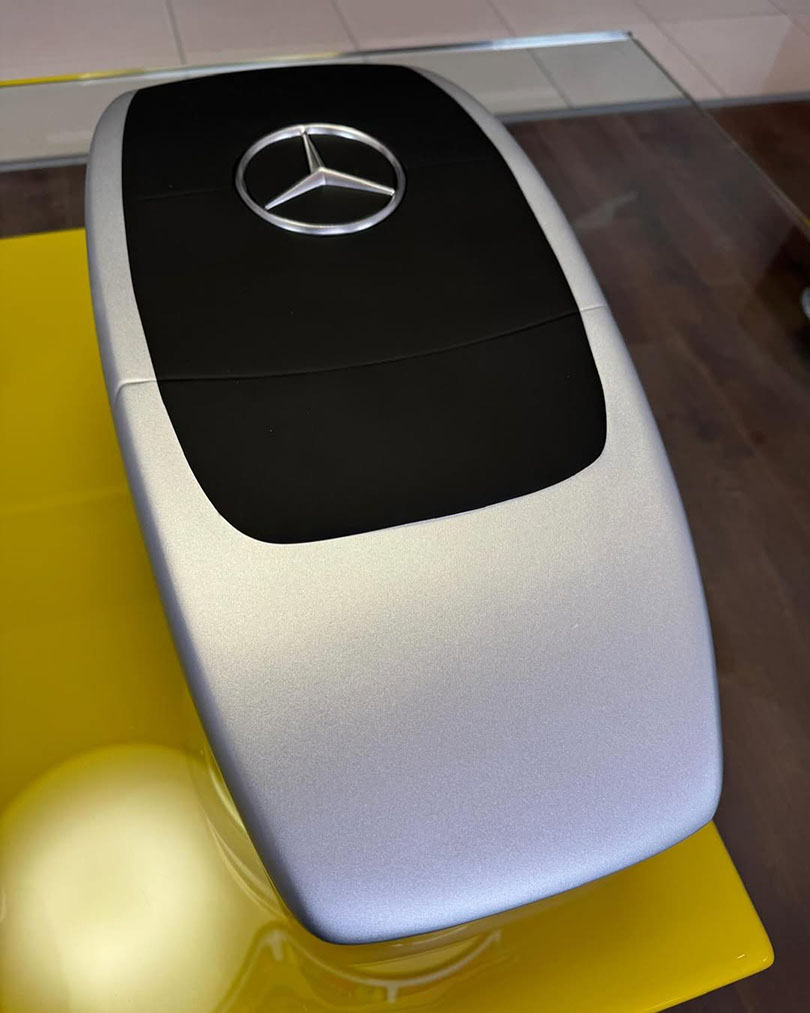 Elegancka figura reklamowa w kształcie pilota Mercedesa, srebrno-czarna figurka z laminatu stojąca na żółtej podstawie.